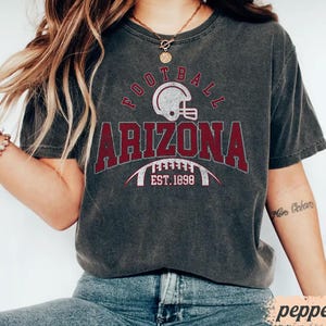 May include: Dark gray t-shirt with the word "FOOTBALL" above "ARIZONA" in maroon letters. A football helmet graphic is above the word "ARIZONA". The text "EST. 1898" is below the word "ARIZONA".