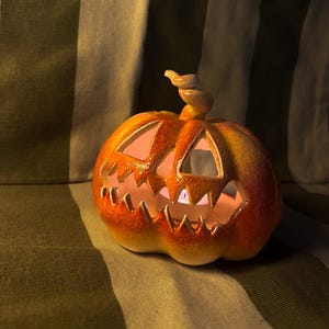 May include: A ceramic jack-o'-lantern with a carved face and a curly stem. The pumpkin is orange with yellow highlights and has triangular eyes and a jagged mouth. The interior glows with a warm light, suggesting a candle inside. A festive Halloween decoration.
