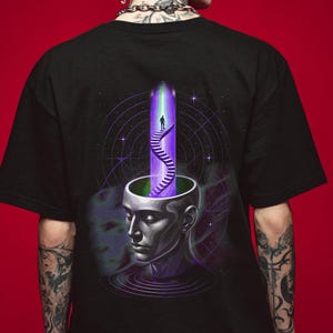 Può includere: T-shirt nero con grafica surreale. Il design raffigura una testa umana con la parte superiore aperta, che rivela una scala che conduce a una luce viola e verde. Una figura sale le scale, su uno sfondo cosmico.