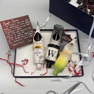 May include: A gift box containing three decorative tattoo machine pen-shaped objects. The pens are white, black, and yellow with decorative designs and text. A handwritten note in Turkish is included, along with red and white ribbon.