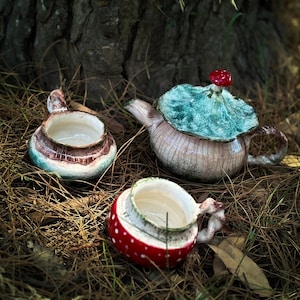 Può includere: Tre pezzi in ceramica a tema fungo: una teiera con coperchio turchese e parte superiore a fungo rosso e due tazze abbinate. I pezzi sono appoggiati su un letto di aghi di pino, con un tronco d'albero sullo sfondo.