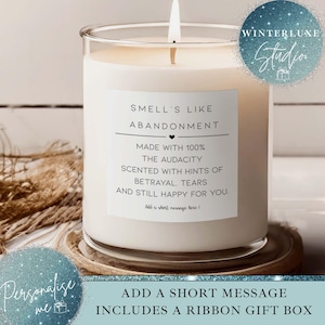 Puede incluir: Una vela blanca perfumada en un tarro de cristal transparente, con una mecha encendida. La etiqueta dice "Smell's Like Abandonment" e incluye el texto "Made with 100% The Audacity, Scented with Hints of Betrayal, Tears and Still Happy for You."