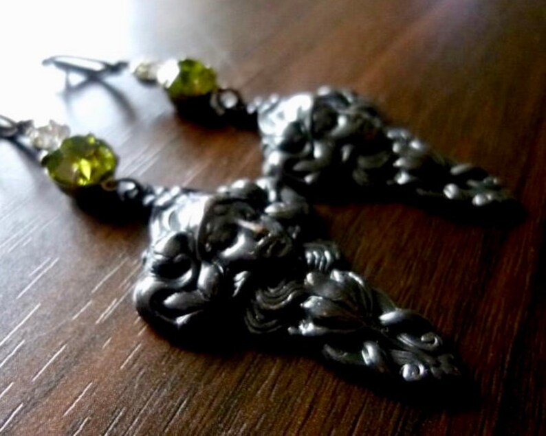 May include: Silver earrings with a detailed face design and green crystal accents.