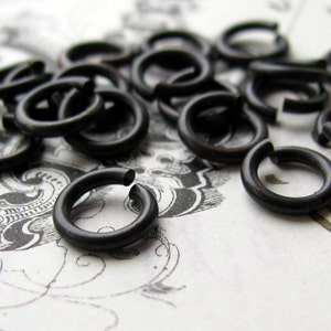 May include: A close-up of a pile of black metal jump rings. The rings are all the same size and are arranged in a random pattern.