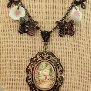 May include: A vintage-inspired necklace with a white rabbit charm, teacups, and butterflies. The necklace is made of metal and has a brown finish.
