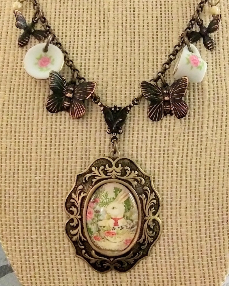 May include: A vintage-inspired necklace with a white rabbit charm, teacups, and butterflies. The necklace is made of metal and has a brown finish.
