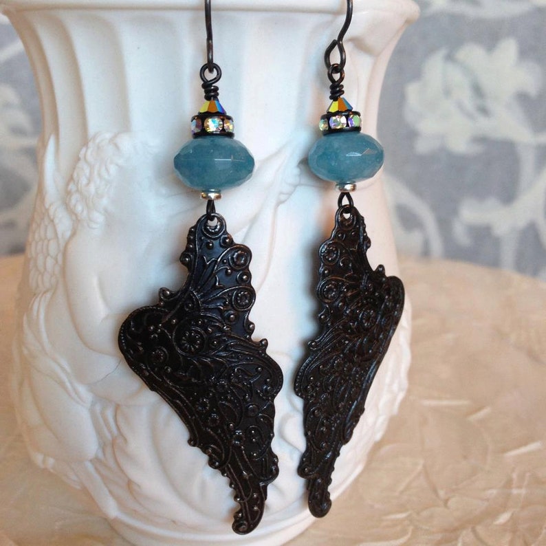 May include: A pair of dangle earrings with a blue gemstone and a black filigree design. The earrings are hanging from a white ceramic vase.