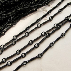 May include: A matte black metal chain featuring a repeating pattern of small circular links connected by elongated, textured bar links. The chain is coiled in a pile at the top left and extends in several parallel strands across the frame.
