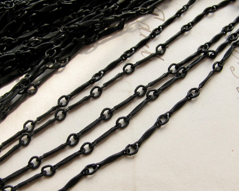 May include: A matte black metal chain featuring a repeating pattern of small circular links connected by elongated, textured bar links. The chain is coiled in a pile at the top left and extends in several parallel strands across the frame.