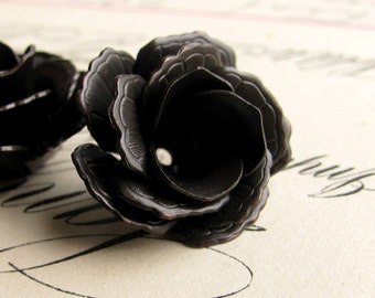 May include: Close-up of two black metal rose-shaped objects. The roses have detailed petal designs and a dark, matte finish. The background is a light, textured surface with faint script.