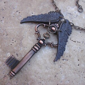 May include: A copper-toned key pendant necklace with a detailed key design and two black wings. The wings are attached to the top of the key and are spread out. The necklace is on a chain.