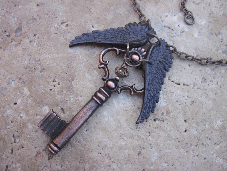 May include: A copper-toned key pendant necklace with a detailed key design and two black wings. The wings are attached to the top of the key and are spread out. The necklace is on a chain.