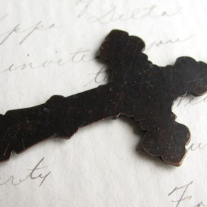 May include: A dark brown metal cross pendant with a decorative, ornate design. The cross has a loop at the top for hanging and is set against a background of handwritten script.