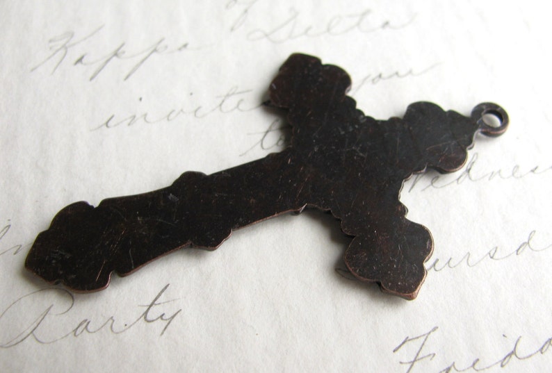 May include: A dark brown metal cross pendant with a decorative, ornate design. The cross has a loop at the top for hanging and is set against a background of handwritten script.