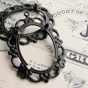 May include: Black metal frame with intricate details, featuring a decorative design with a floral motif. The frame is oval shaped and has a small loop at the top.