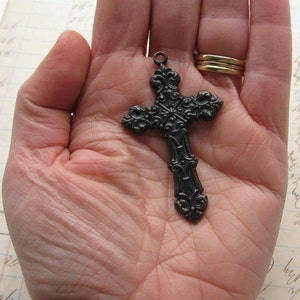 May include: Ornate, dark metal cross pendant with intricate scrollwork and a loop for a chain. The cross is held in an open hand, showcasing its detailed design. A gold ring is visible in the background.