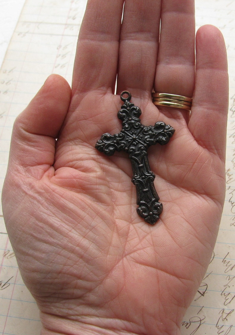 May include: Ornate, dark metal cross pendant with intricate scrollwork and a loop for a chain. The cross is held in an open hand, showcasing its detailed design. A gold ring is visible in the background.