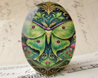 May include: An oval glass cabochon featuring a vibrant green luna moth centered against a dark blue background with intricate gold filigree patterns and small pink floral accents. The surface is smooth and glossy with a domed, reflective finish.