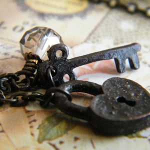 Op de afbeelding: Close-up van een vintage-stijl ketting met een kleine, donkere metalen sleutel, een hartvormig hangslot en een facetgeslepen kristallen kraal. De ketting en bedels hebben een rustieke, antieke afwerking, wat een romantisch of gotisch thema suggereert.