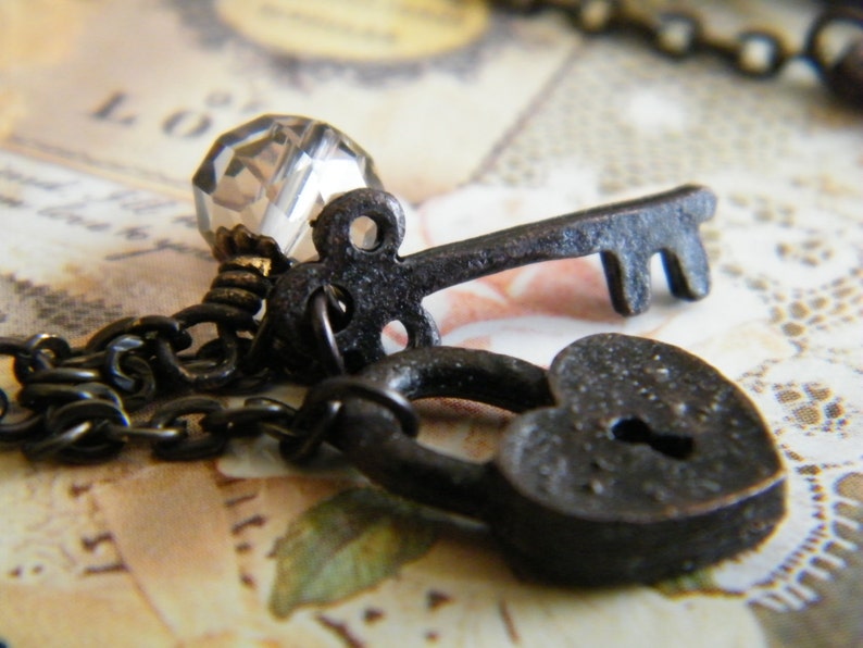 Op de afbeelding: Close-up van een vintage-stijl ketting met een kleine, donkere metalen sleutel, een hartvormig hangslot en een facetgeslepen kristallen kraal. De ketting en bedels hebben een rustieke, antieke afwerking, wat een romantisch of gotisch thema suggereert.
