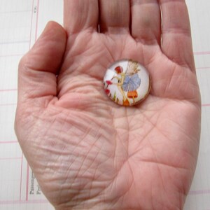 May include: A glass cabochon with a fairy illustration. The fairy is wearing a blue dress and is holding a flower. The cabochon is oval shaped and has a clear background.