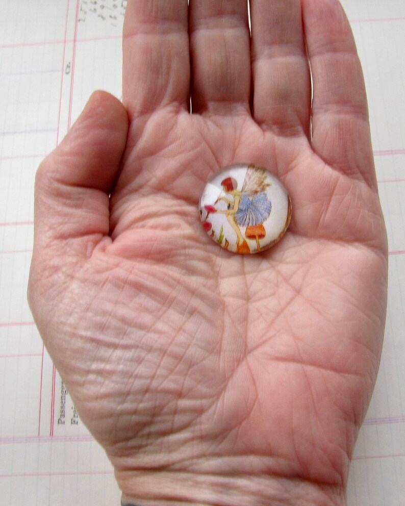May include: A glass cabochon with a fairy illustration. The fairy is wearing a blue dress and is holding a flower. The cabochon is oval shaped and has a clear background.