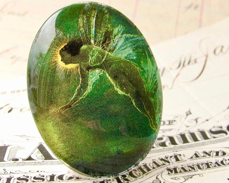 Op de afbeelding: Een groene glazen cabochon met een vintage afbeelding van een fee met vleugels, die vliegt met een gloeiende halo. De fee is afgebeeld in een groen-wit kleurenschema.