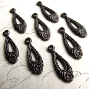 May include: Close-up of eight antique bronze teardrop charms. Each charm features an open teardrop shape with decorative detailing at the top and bottom. The charms are arranged on a light-colored surface.