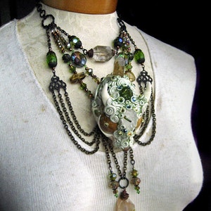 Puede incluir: Un collar de m&uacute;ltiples cadenas con un colgante de calavera de cer&aacute;mica blanca adornado con cristales verdes y transparentes, cadenas marrones y cuentas verdes y marrones. El collar se muestra en un maniqu&iacute; blanco.