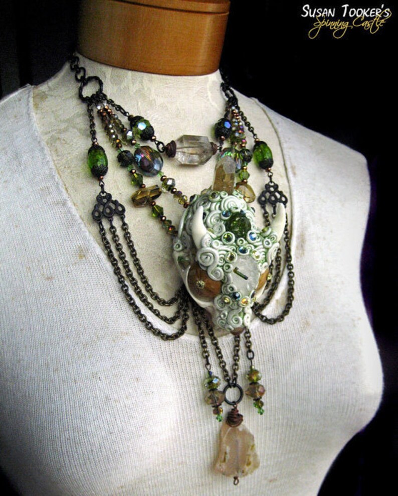 Puede incluir: Un collar de m&uacute;ltiples cadenas con un colgante de calavera de cer&aacute;mica blanca adornado con cristales verdes y transparentes, cadenas marrones y cuentas verdes y marrones. El collar se muestra en un maniqu&iacute; blanco.