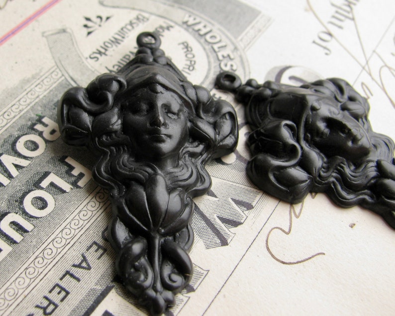 May include: Two black resin decorative embellishments with a woman's face and flowing hair. The embellishments are in the shape of a woman's head with a floral design.