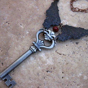 May include: A silver key with a decorative ornate head and a chain with two black metal wings attached to it. A red gemstone is visible in the center of the wings.
