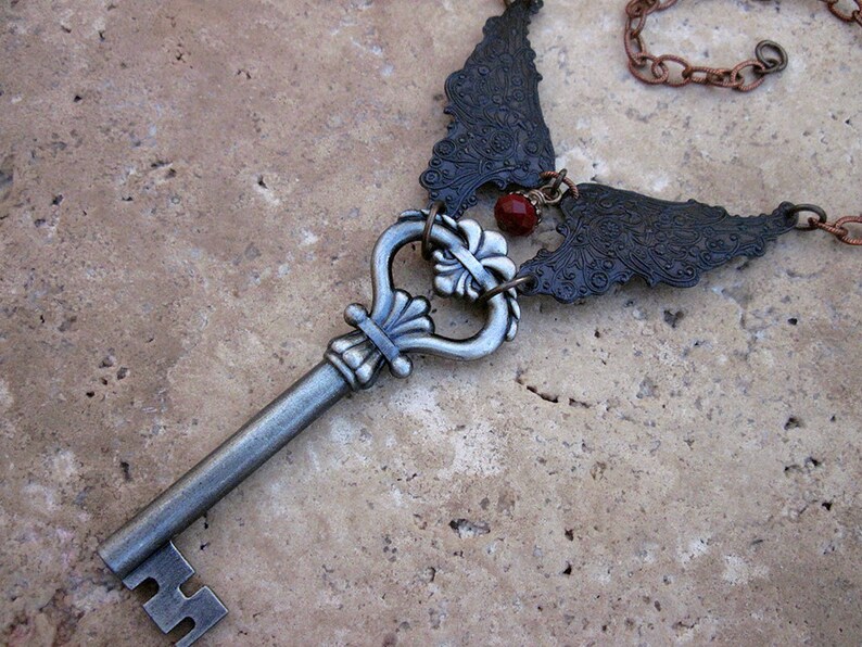 May include: A silver key with a decorative ornate head and a chain with two black metal wings attached to it. A red gemstone is visible in the center of the wings.