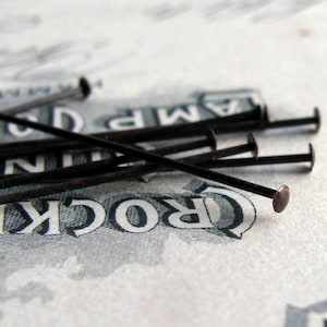 May include: A close-up of a group of black sewing pins with silver heads. The pins are arranged in a pile on a white surface with the word "CROCKER" printed on it.