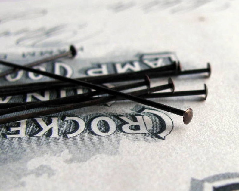 May include: A close-up of a group of black sewing pins with silver heads. The pins are arranged in a pile on a white surface with the word "CROCKER" printed on it.