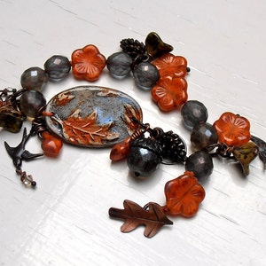 May include: A bracelet with orange and gray beads, a ceramic charm with a leaf design, and a small acorn charm. The bracelet has a rustic, autumnal feel.