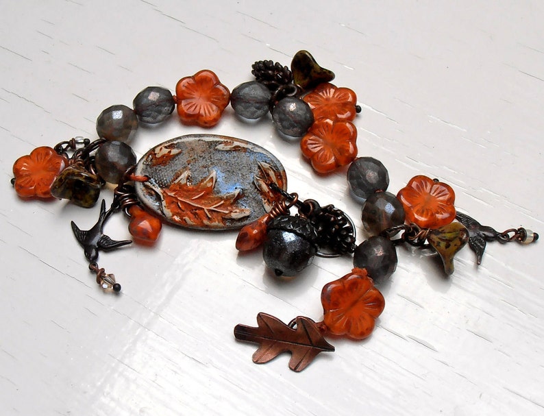 May include: A bracelet with orange and gray beads, a ceramic charm with a leaf design, and a small acorn charm. The bracelet has a rustic, autumnal feel.