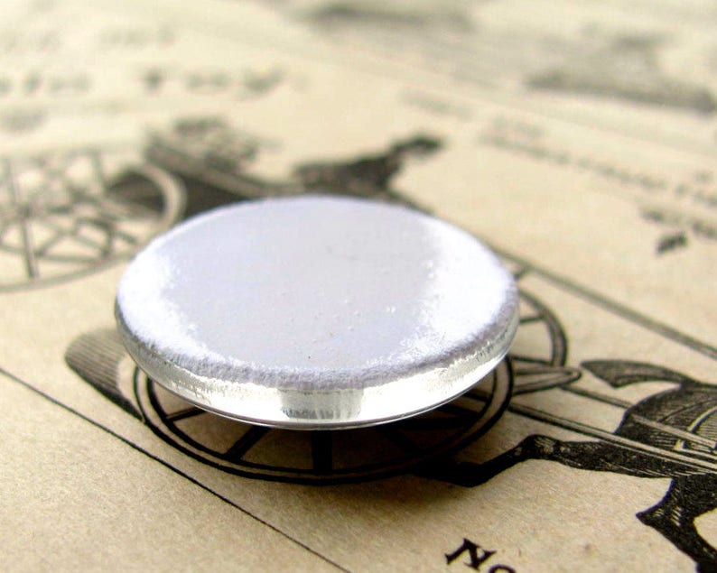 May include: A white round button with a silver metal rim. The button is blank and ready to be customized.