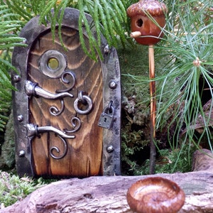 K&ouml;nnte beinhalten: Eine Miniatur-Feent&uuml;r aus Holz mit Metallakzenten, ein kleiner Eichel-Vogelhaus und ein Pilz befinden sich inmitten von Gr&uuml;n. Die T&uuml;r hat einen Griff, einen Ring und ein Vorh&auml;ngeschloss. Das Eichel-Vogelhaus ist auf einem Holzpfahl montiert.