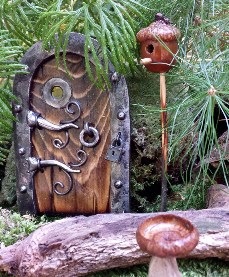 K&ouml;nnte beinhalten: Eine Miniatur-Feent&uuml;r aus Holz mit Metallakzenten, ein kleiner Eichel-Vogelhaus und ein Pilz befinden sich inmitten von Gr&uuml;n. Die T&uuml;r hat einen Griff, einen Ring und ein Vorh&auml;ngeschloss. Das Eichel-Vogelhaus ist auf einem Holzpfahl montiert.
