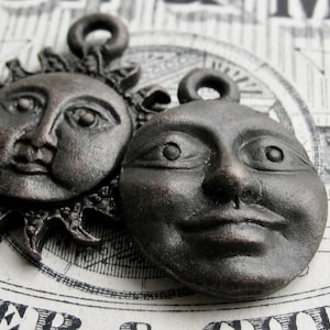 May include: Two antique-style metal charms, one depicting a sun with a face and the other a moon with a face. Both charms are in a dark bronze color.