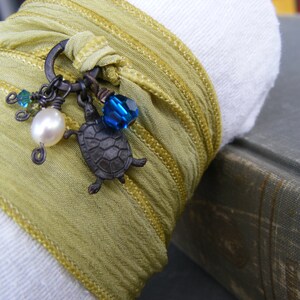 May include: A green silk ribbon bracelet with a charm featuring a turtle, a pearl, and blue glass beads.