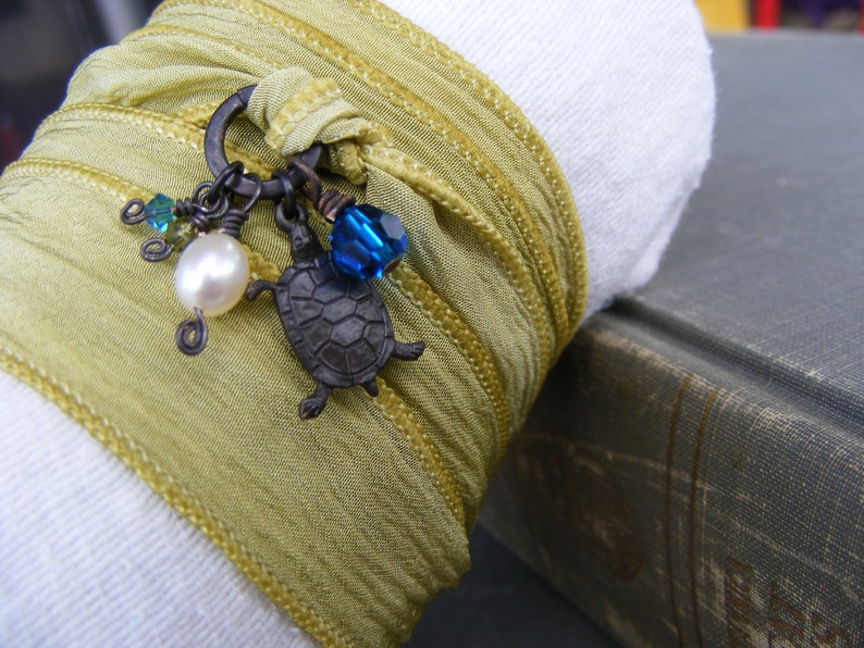 May include: A green silk ribbon bracelet with a charm featuring a turtle, a pearl, and blue glass beads.