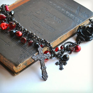 May include: A gothic-style necklace with a dark chain, featuring red beads, skull charms, and ornate crosses. A black rose pendant and an antique book with the text "ELEMENTS OF NATURAL PHILOSOPHY" are also visible.