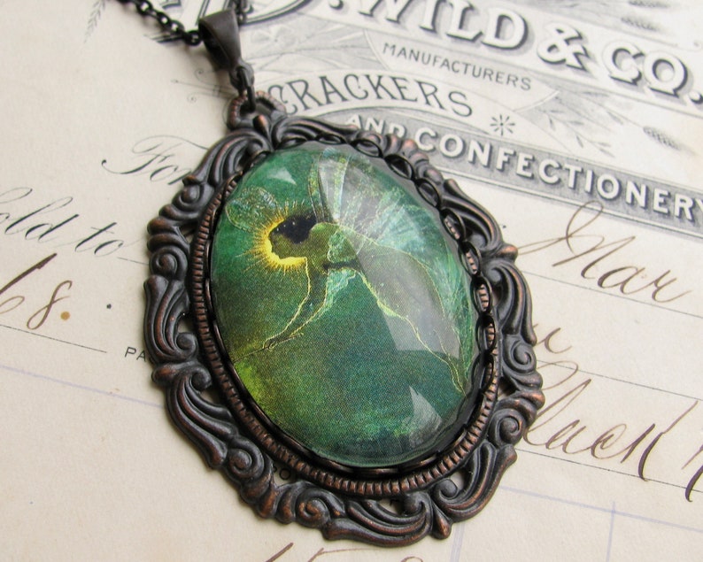 Op de afbeelding: Een bronzen hanger met een sierlijk frame en een groene glazen cameo met een fee met gloeiende vleugels.