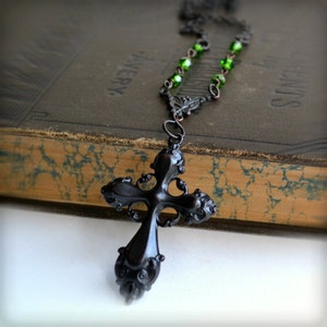 May include: A black metal cross pendant with intricate details hangs from a necklace with green glass beads. The necklace is resting on a vintage book.