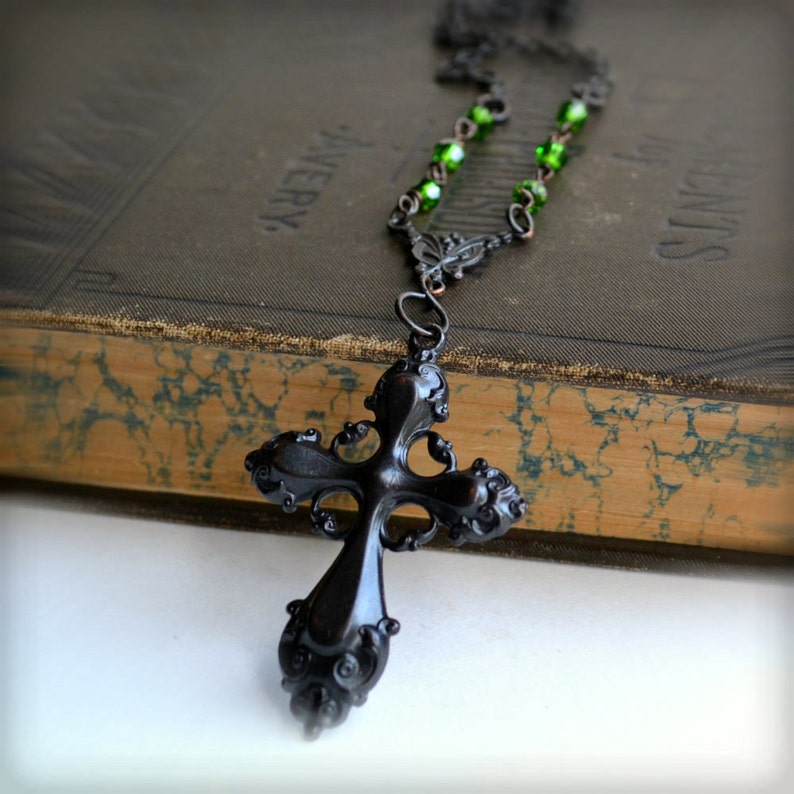 May include: A black metal cross pendant with intricate details hangs from a necklace with green glass beads. The necklace is resting on a vintage book.