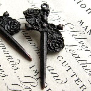 May include: Two black metal sword charms with floral details. The swords have a pointed tip and are decorated with roses and leaves.