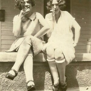 May include: Two women in vintage clothing are sitting on a porch, each holding a cigarette. They are both wearing white socks and dark shoes. The woman on the left is wearing a white dress with a collar and a dark skirt. The woman on the right is wearing a white dress with short sleeves.