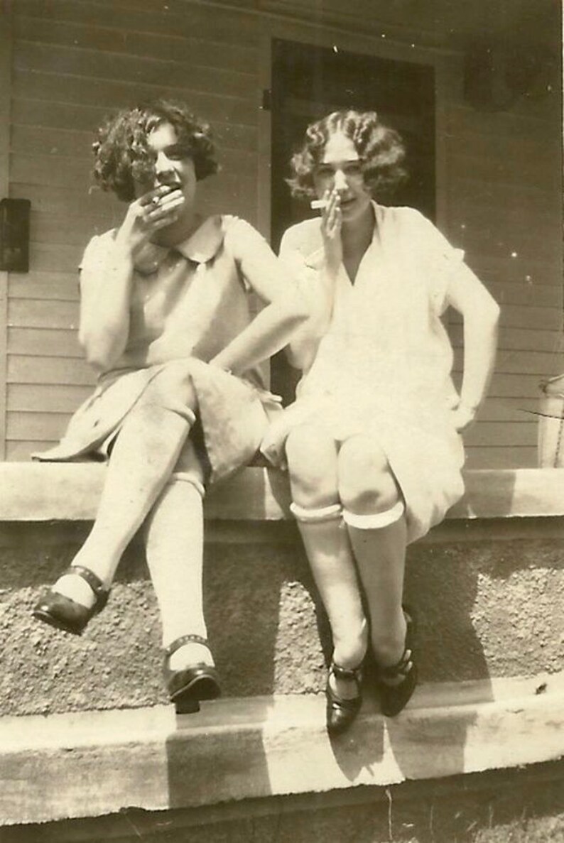 May include: Two women in vintage clothing are sitting on a porch, each holding a cigarette. They are both wearing white socks and dark shoes. The woman on the left is wearing a white dress with a collar and a dark skirt. The woman on the right is wearing a white dress with short sleeves.